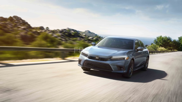 Front view of a Honda Civic Sedan as it drives past a rocky shoreline full of greenery.