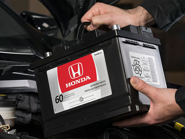 Close-up shot of Honda technician inserting a genuine Honda battery into an open car hood.  	Gros plan d’un technicien Honda insérant une véritable batterie Honda dans un capot de voiture ouvert.