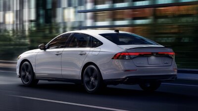 3/4 rear view of white Accord driving on city side highway past buildings at night. 