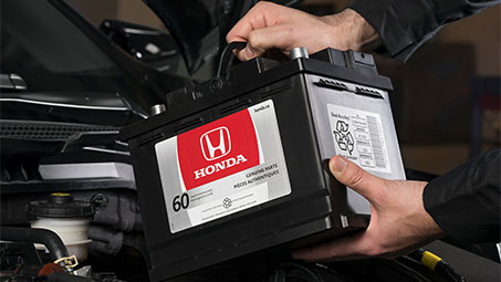 Image of a Honda technician reaching into an open hood to maintain and inspect the condition of a genuine Honda battery and charging system. // Image d'un technicien Honda travaillant dans un capot ouvert pour entretenir et inspecter l'état d'une batterie et d'un système de charge Honda d'origine.