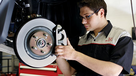 Honda technician performing maintenance on a Honda brake system’s hydraulic component while holding the brake pad and closely examining it. // Technicien Honda effectuant l'entretien d'un composant hydraulique du système de freinage Honda tout en tenant la plaquette de frein et en l'examinant de près.