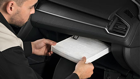 A Honda technician reaching to install and inspect an air filter in the cabin of a Honda, with black interior finishes. // Un technicien Honda s’approche pour installer et inspecter un filtre à air dans l'habitacle d'une Honda, avec des finitions intérieures noires.