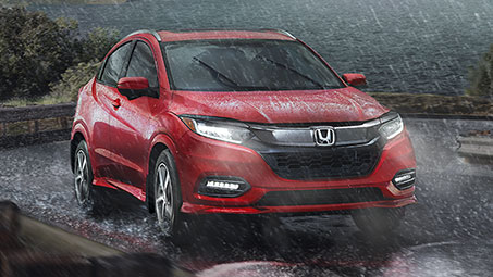 Front-view of a red Honda SUV driving on a road with headlights on, through rainy and stormy weather conditions. 	Vue de face d'un VUS Honda rouge roulant sur une route avec les phares allumés, par temps de pluie et d'orage.