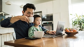 A father drinks from a mug in front of his laptop while his son sits on his lap. / Un père portant une tasse à ses lèvres devant son ordinateur portable, son fils assis sur ses genoux.