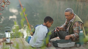 A grandfather and grandson sit by a lake fishing together. / Un grand-père et son petit-fils pêchant ensemble assis au bord d’un lac.