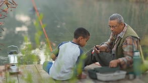 A grandfather and grandson sit by a lake fishing together. / Un grand-père et son petit-fils pêchant ensemble assis au bord d’un lac. 