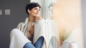 A young woman sits wrapped in a blanket looking out the window. / Une jeune femme, assise avec une couverture, regardant par la fenêtre.
