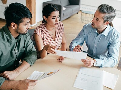 Three people sit around a table discussing paperwork. / Trois personnes sont assises autour d'une table pour discuter au sujet de documents.