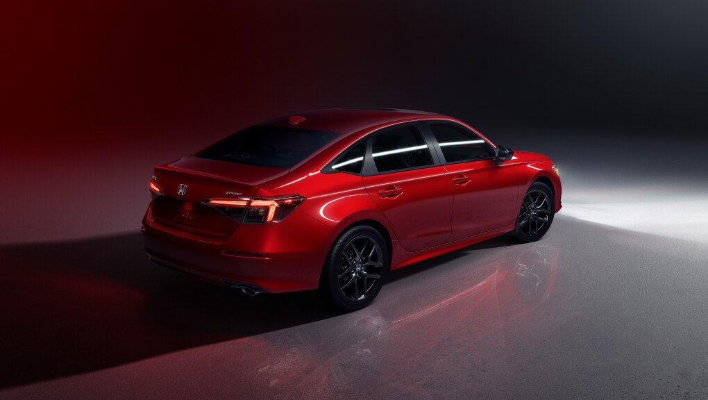 Vue latérale arrière d’une Honda Civic 2022 rouge dans un garage sombre, le rouge du véhicule se reflète sur le sol et les murs.
