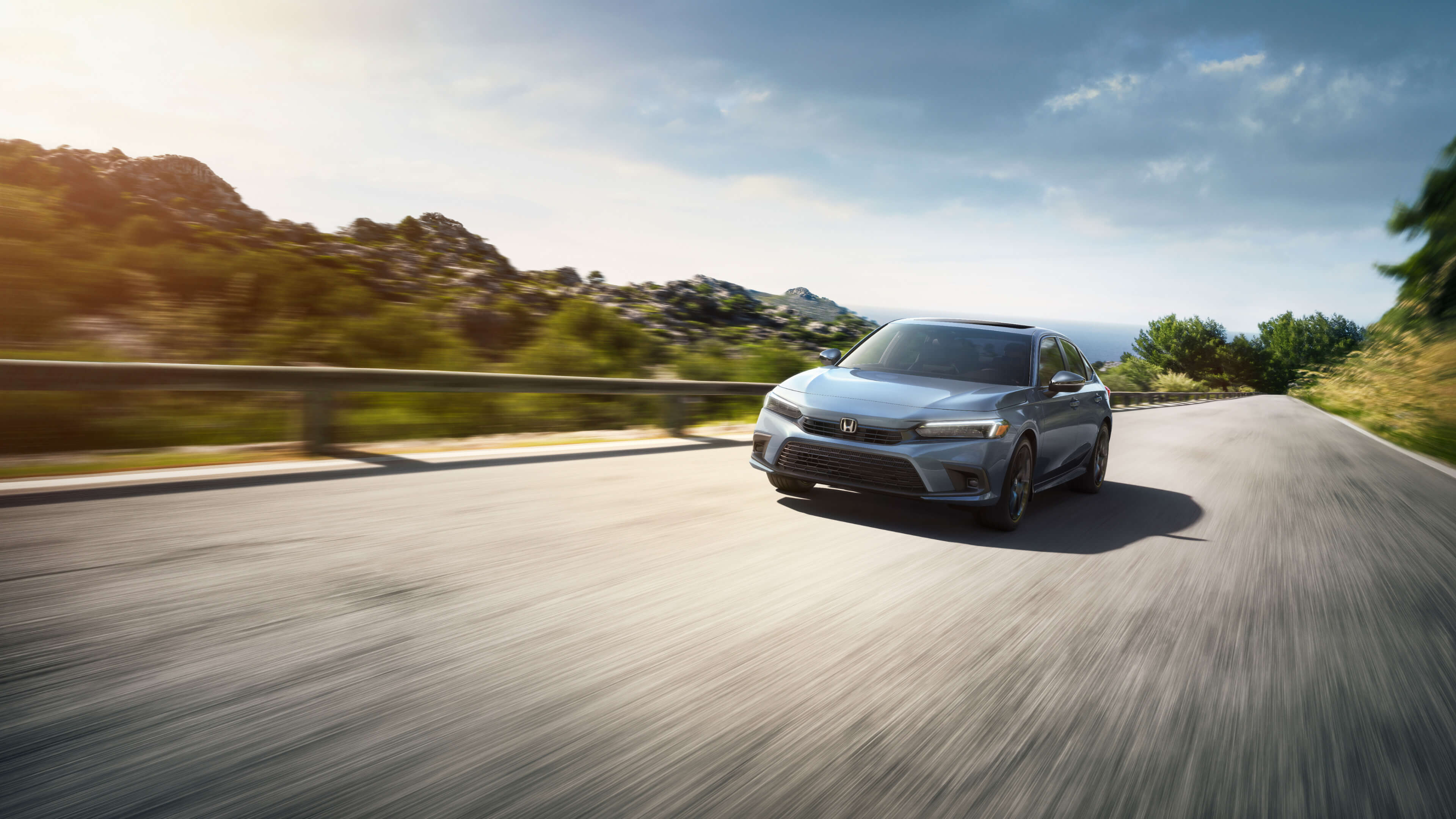 A grey 2022 Honda Civic blurred by motion as it drives on a countryside road with rolling hills in the background.