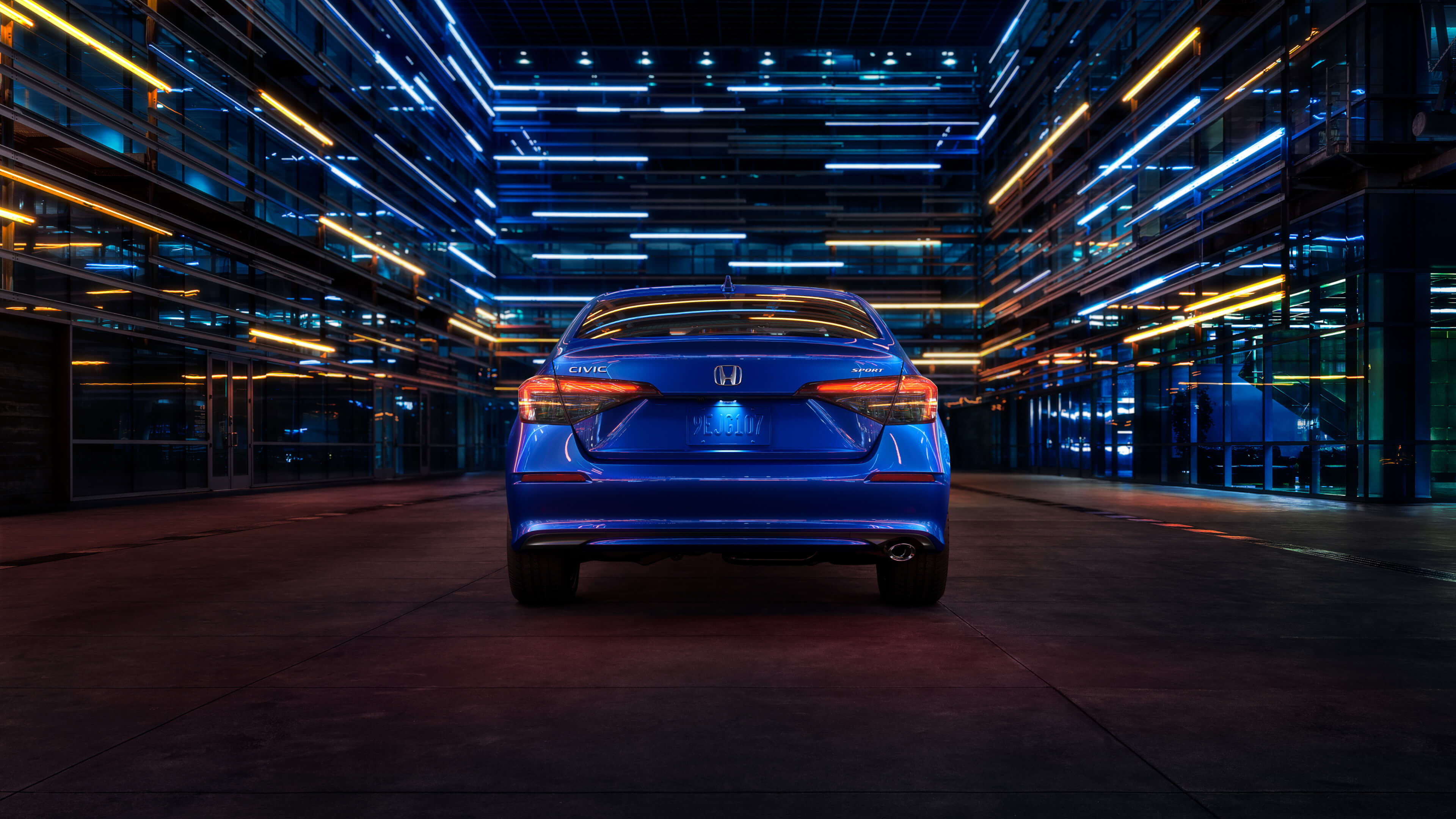 Rear view of a blue 2022 Honda Civic driving at night through a city street illuminated by blue and white lights emanating from the surrounding buildings.