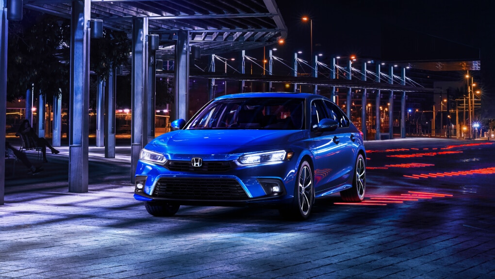 Front end, side-angled view of a blue 2022 Honda Civic driving past a row of pillars at night.