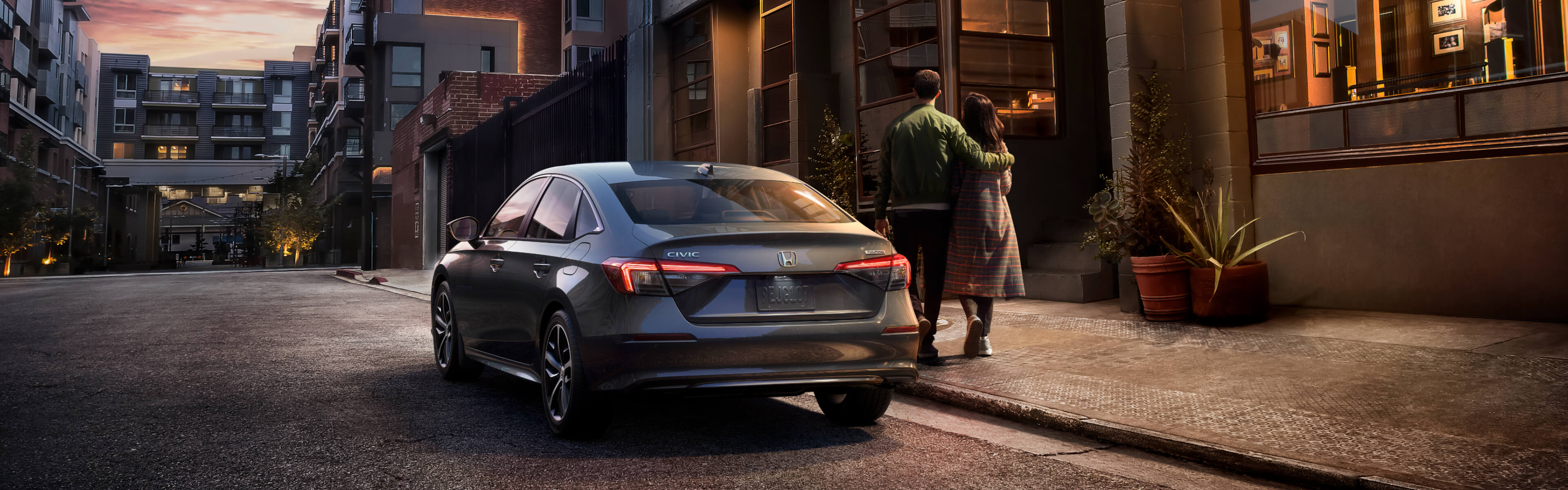 Vue arrière d’une Honda Civic 2022 grise stationnée dans un quartier résidentiel en début de soirée, avec un couple enlacé qui s’éloigne.