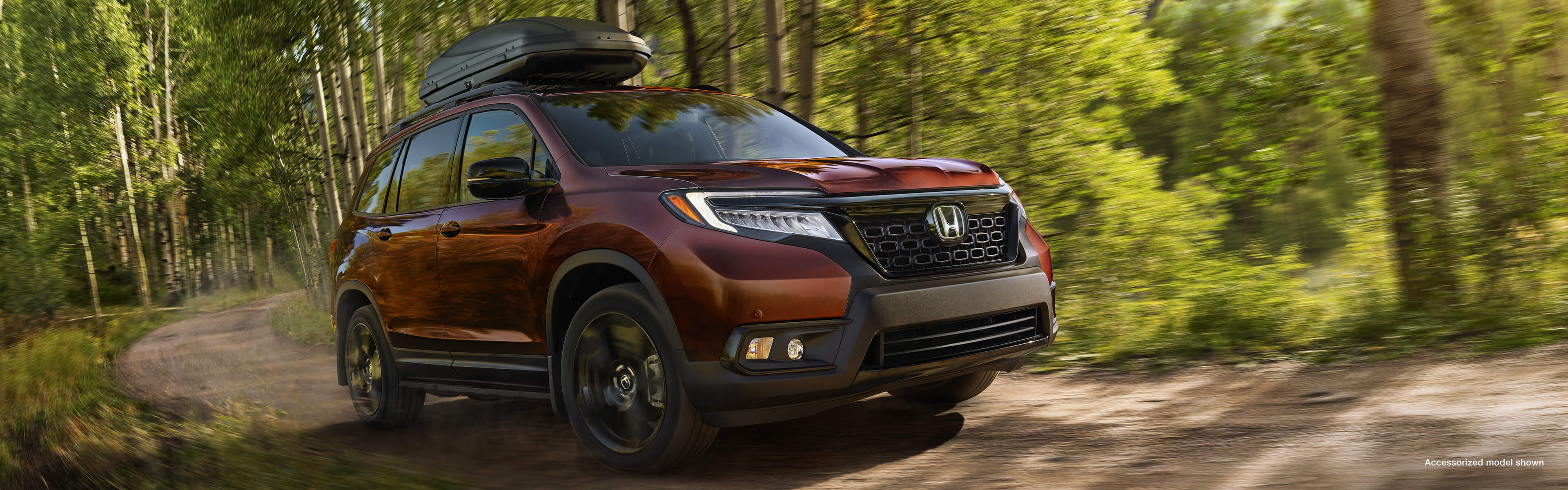 Front 3/4 view of 2021 Honda Passport Touring in Deep Scarlet Pearl with Honda Genuine Accessories, driving on a forest road.