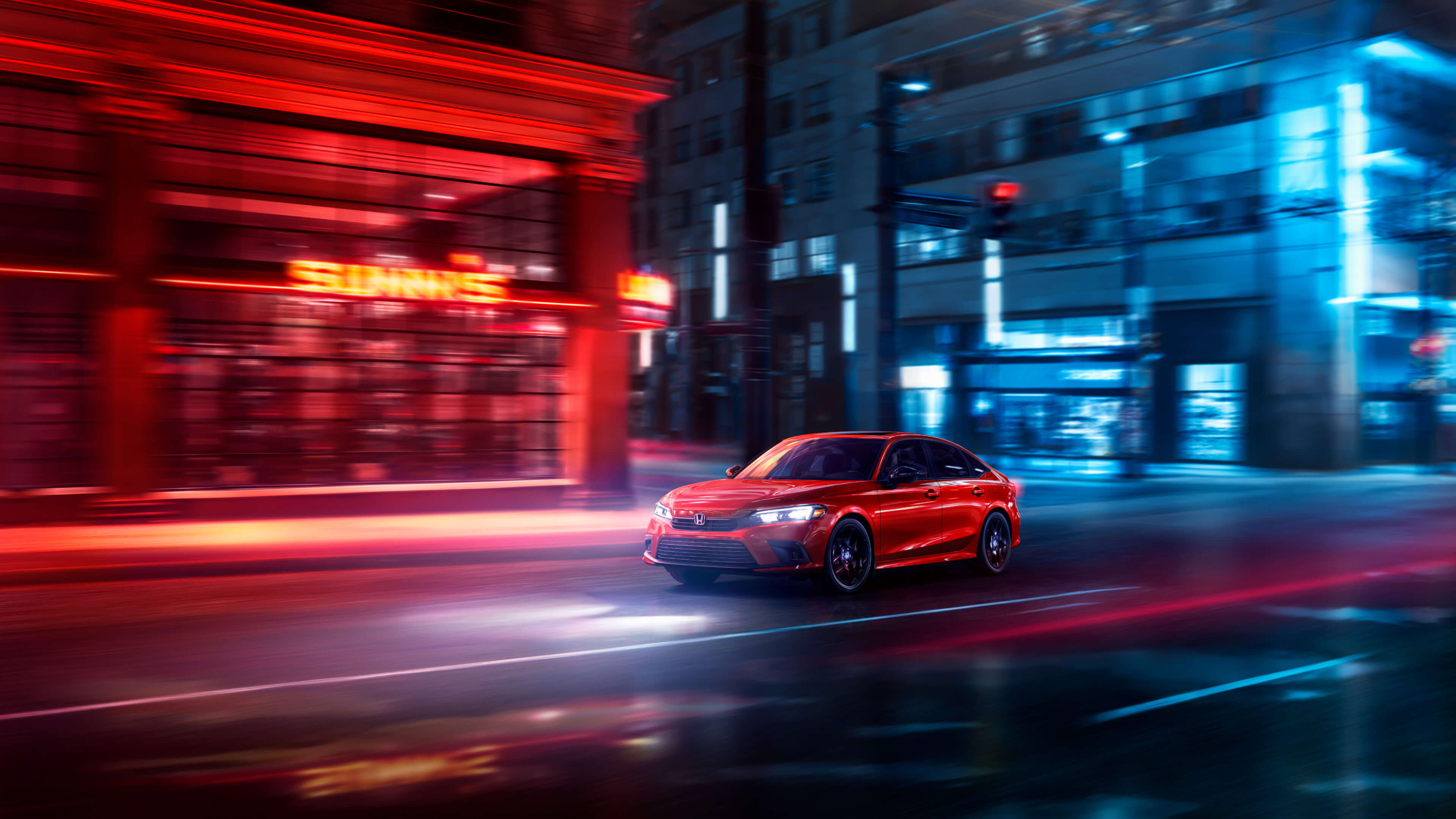 Vue latérale avant d’une Honda Civic 2022 rouge traversant la ville de nuit. L’image est floue, pour rendre l’impression de mouvement, et éclairée par des lumières rouges et bleues émises par les bâtiments de la rue.