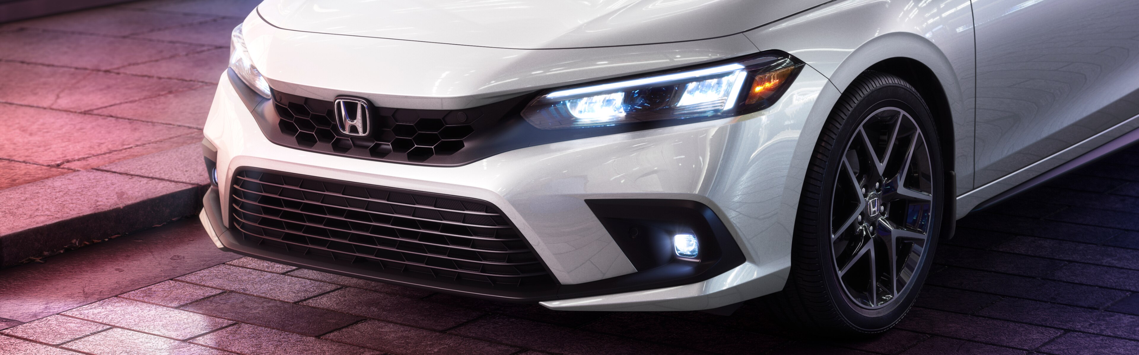 Side-angled close-up of white Honda Civic Hatchback with headlights shining on a brick road
.