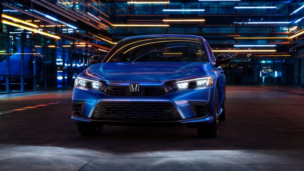 Front view of a blue 2022 Honda Civic driving at night through a city street illuminated by blue and white lights emanating from the buildings.