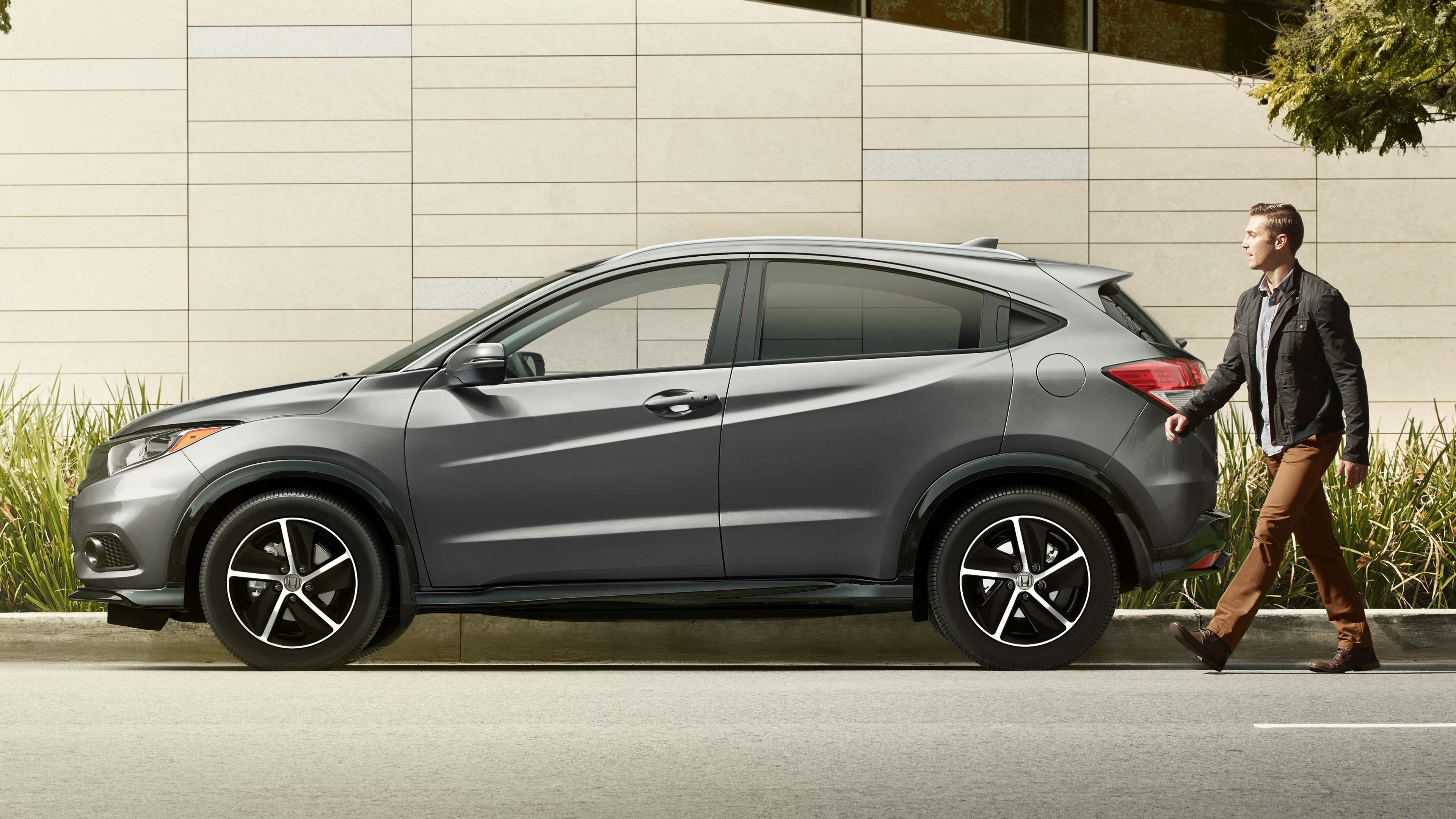 Driver-side profile view of the 2022 Honda HR-V Sport in silver parked in front of a modern building, with a man walking up from behind. 