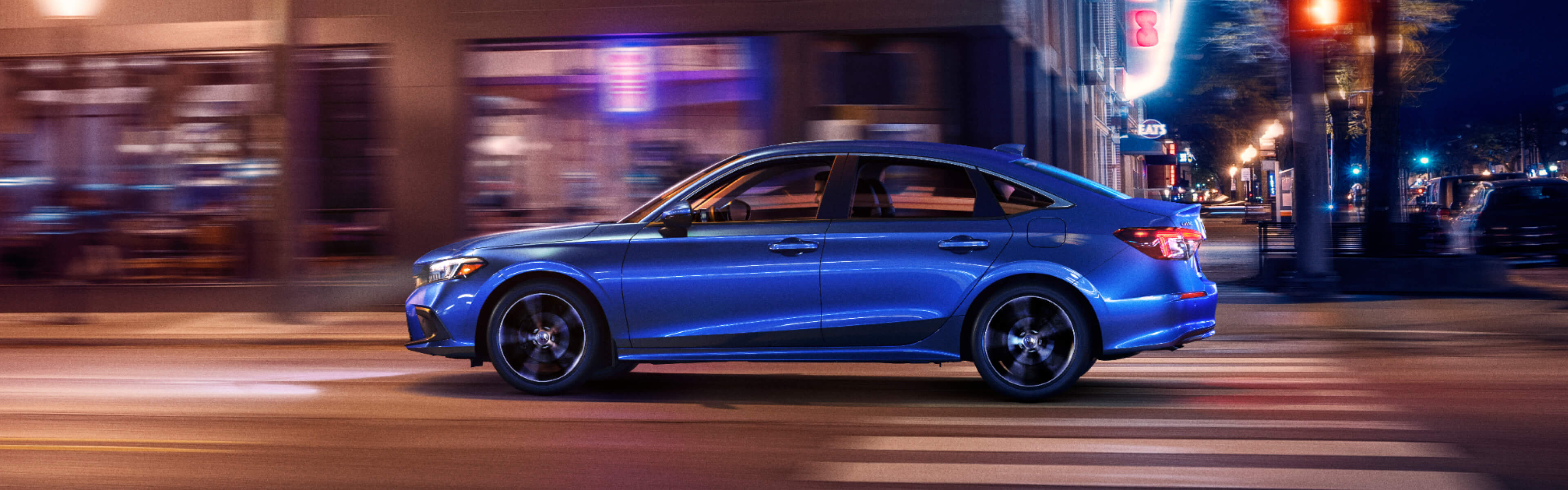 Vue latérale d’une Honda Civic bleue 2022 circulant de nuit dans un centre-ville. L’image est légèrement floue pour rendre le mouvement de la prise de vue.