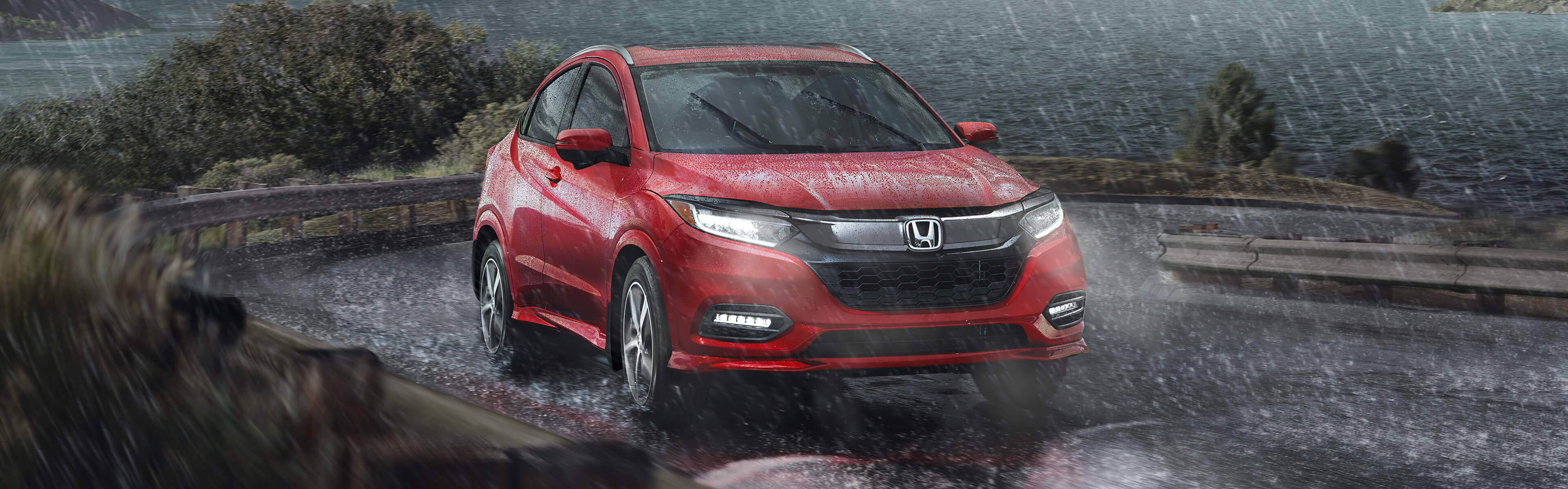 Front passenger-side view of the 2022 Honda HR-V in red driving on lakeside road in the rain. 