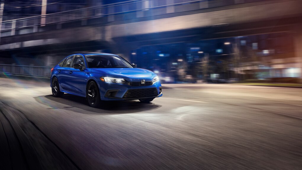 Front passenger-side view of a blue 2022 Honda Civic seen driving at night past an overpass with the city blurred in the background.