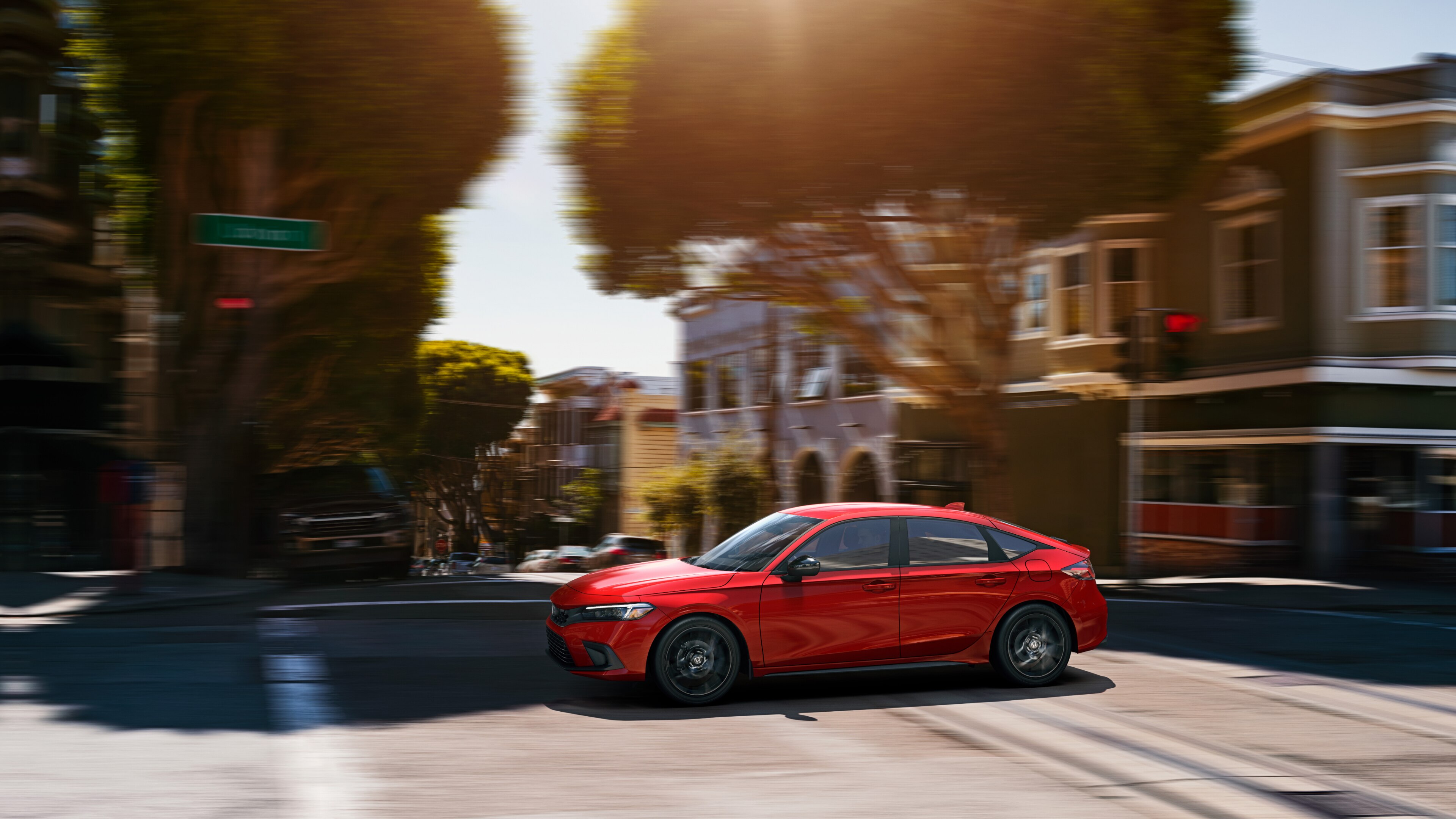 Side view of red Honda Civic Hatchback driving through an intersection with trees and houses in the background